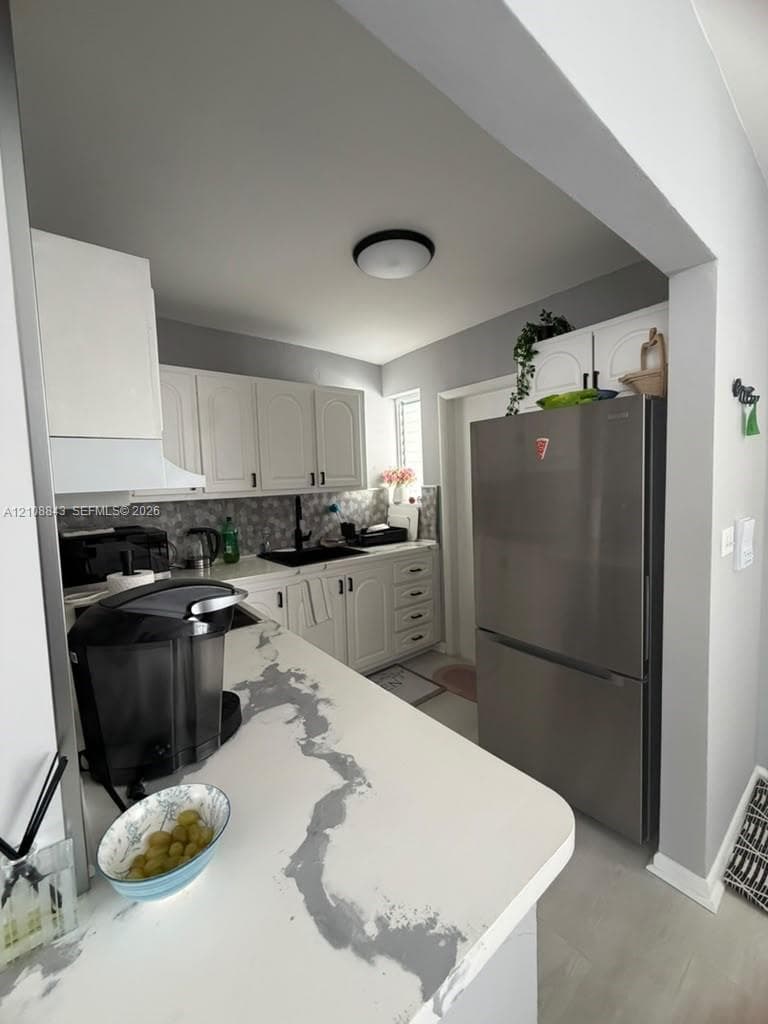 Kitchen featuring white cabinetry, stone-patterned countertops, a stainless steel refrigerator, patterned tile backsplash, and a black faucet