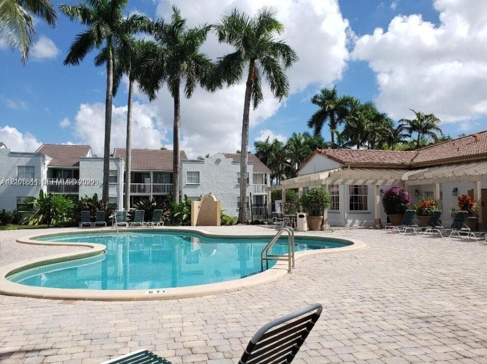 Free-form swimming pool with metal ladder entry, surrounded by expansive paver patio, wood-beam pergola, and established palm trees