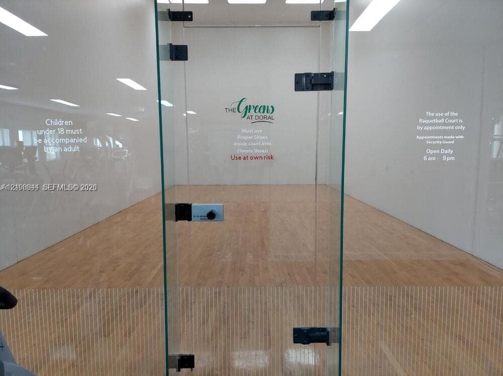 Indoor racquetball court featuring wood-finish flooring, high-gloss white walls, and a glass-enclosed entry