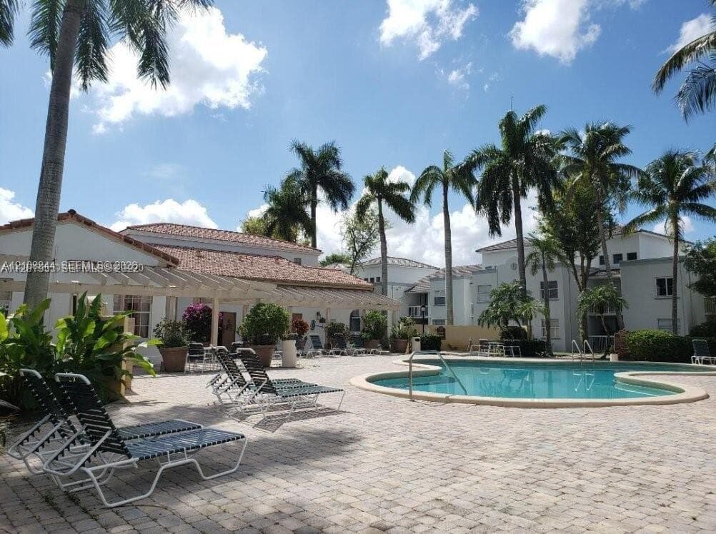 Outdoor swimming pool with curved perimeter, surrounding paver patio, and attached wooden trellis structure amidst mature palm trees