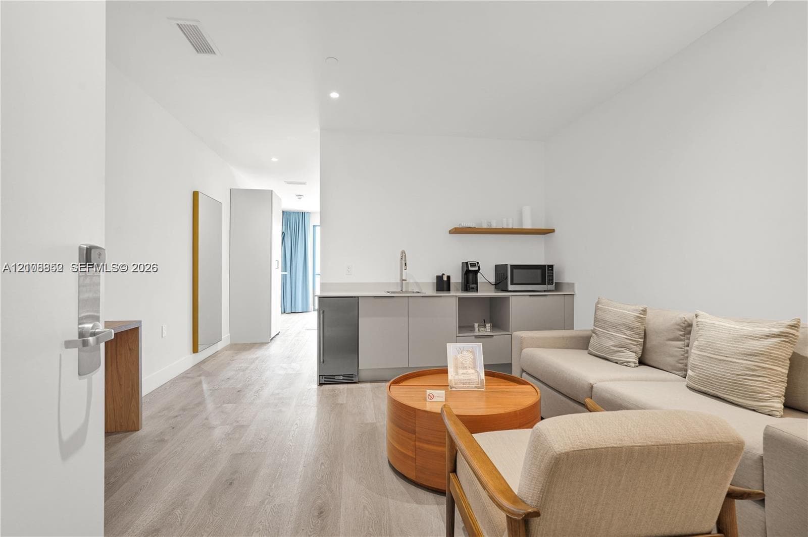 Kitchenette featuring matte-finish cabinetry, stainless steel under-counter refrigerator, sink with faucet, and floating wood shelf, complemented by wood-finish flooring
