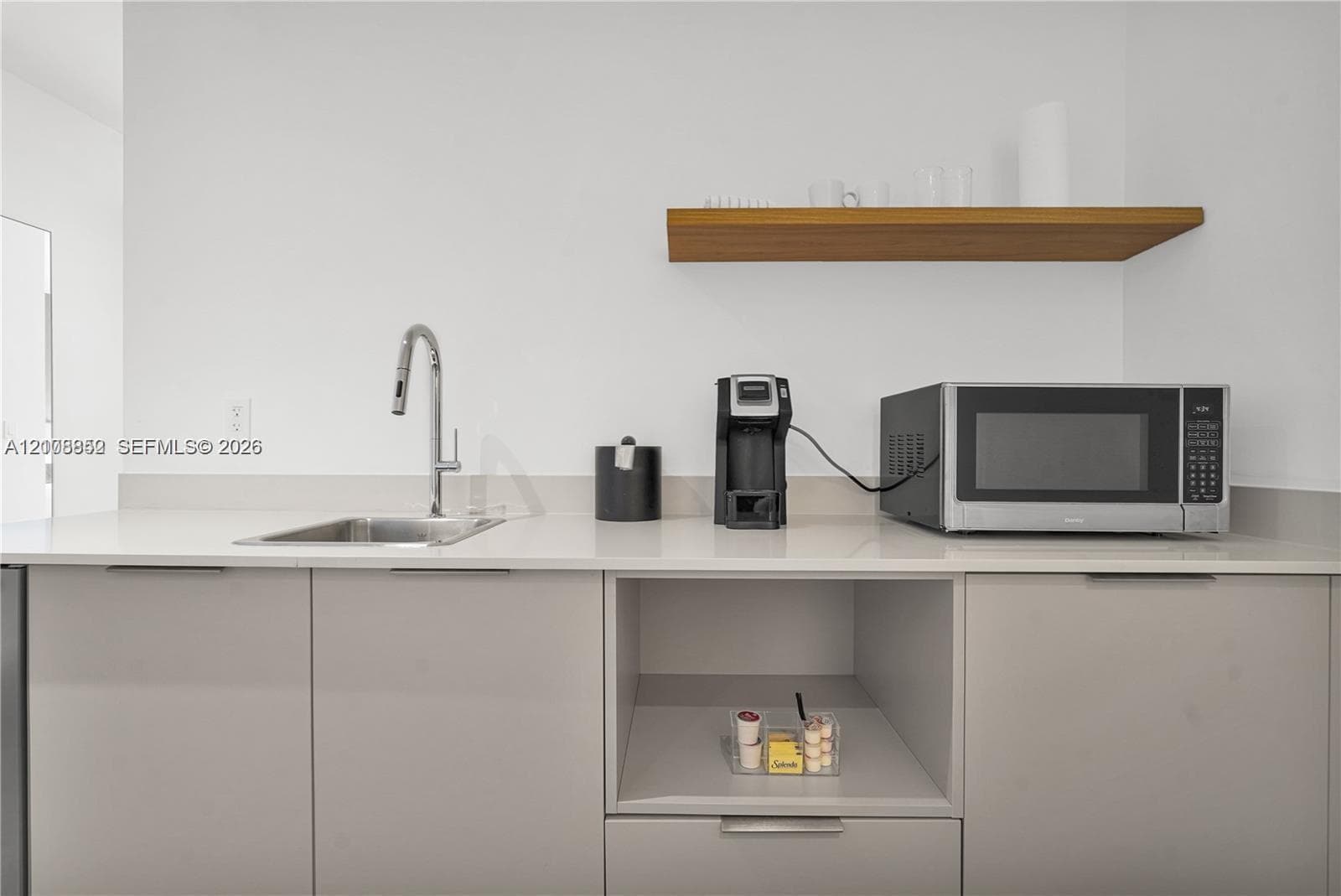 Modern kitchenette featuring flat-panel cabinetry, light-toned countertop, stainless steel undermount sink with gooseneck faucet, and floating wood-finish shelving