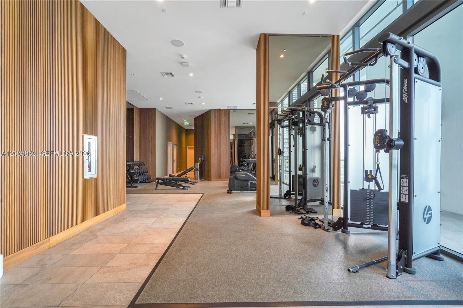 Fitness facility featuring vertical wood paneling, floor-to-ceiling windows, mirrored structural columns, and tiled flooring