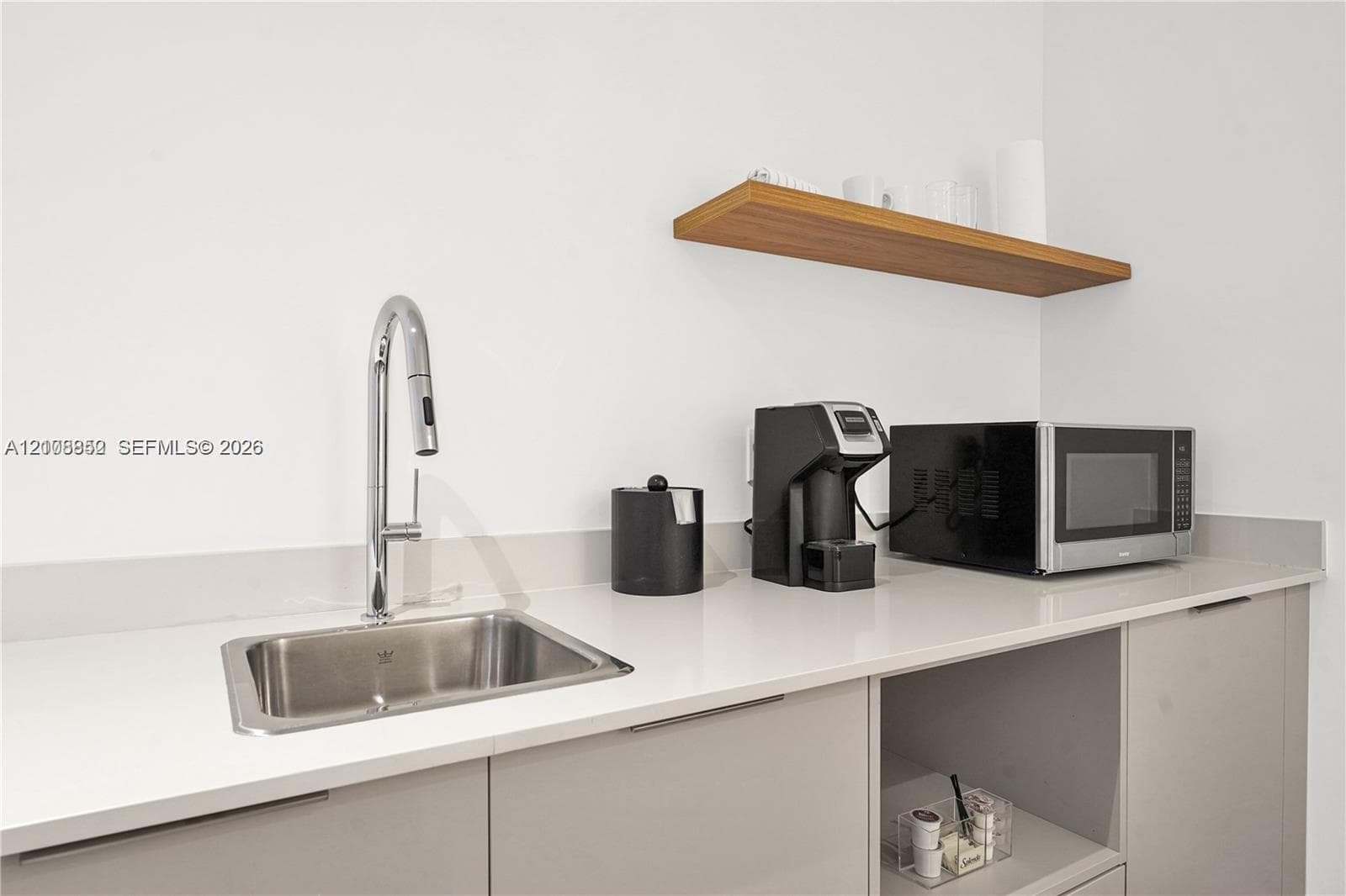 Wet bar area featuring flat-panel cabinetry, light-toned countertop with matching backsplash, undermount stainless steel sink, pull-down chrome faucet, and wood-finish wall-mounted shelving