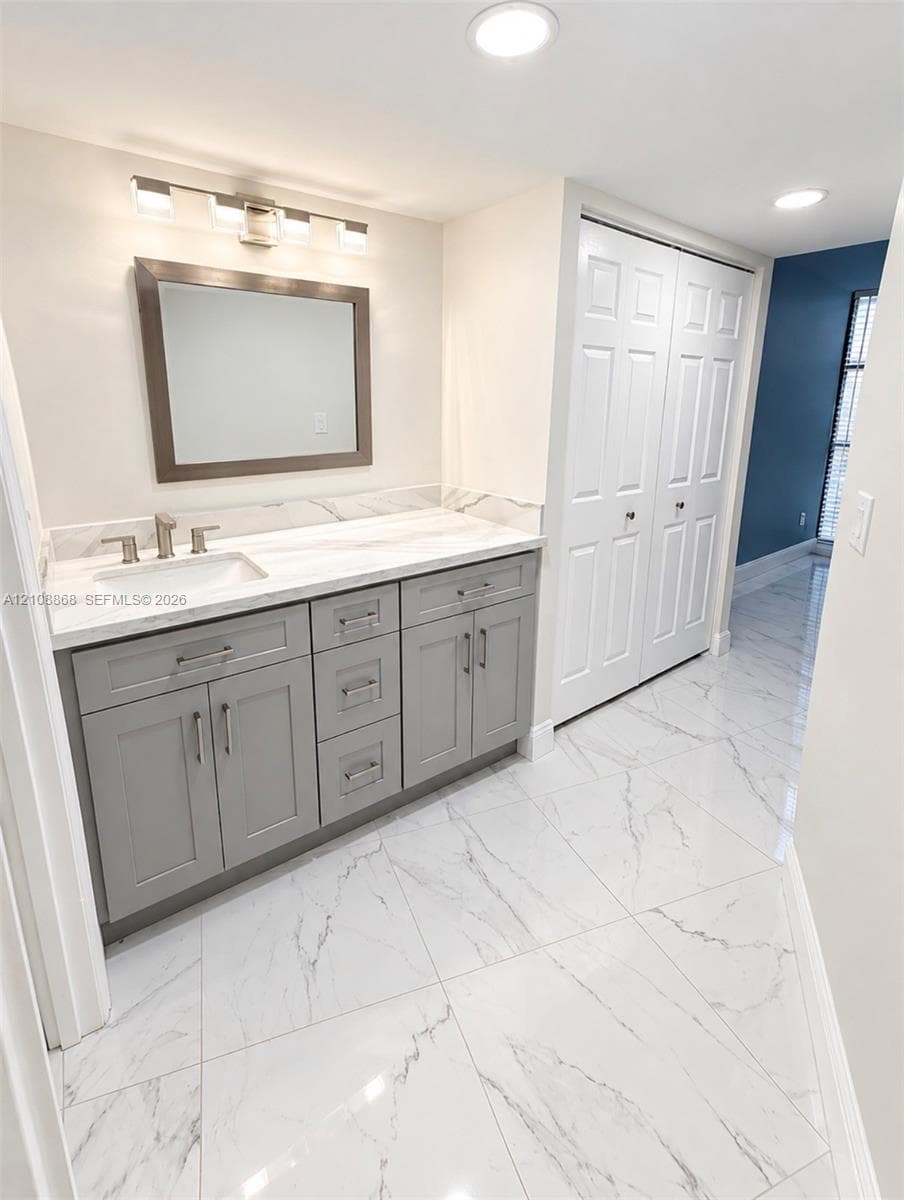 Grey shaker-style vanity featuring a stone-finish countertop, rectangular undermount sink, dual-handle faucet, and large-format porcelain tile flooring