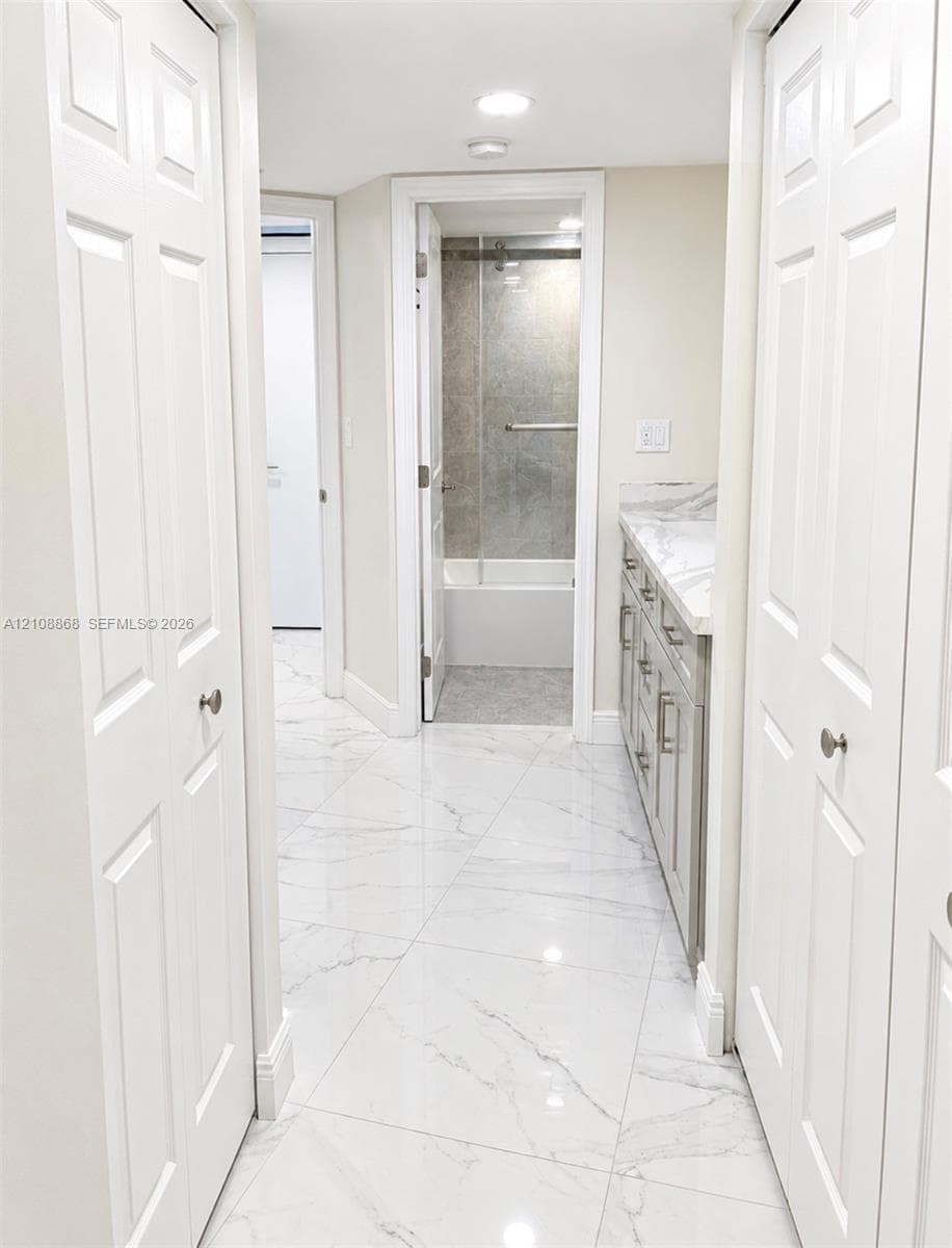 Bathroom featuring large-format stone-finish flooring, a vanity with stone-finish countertop, dual bi-fold closet doors, and a shower-tub combination with a glass enclosure