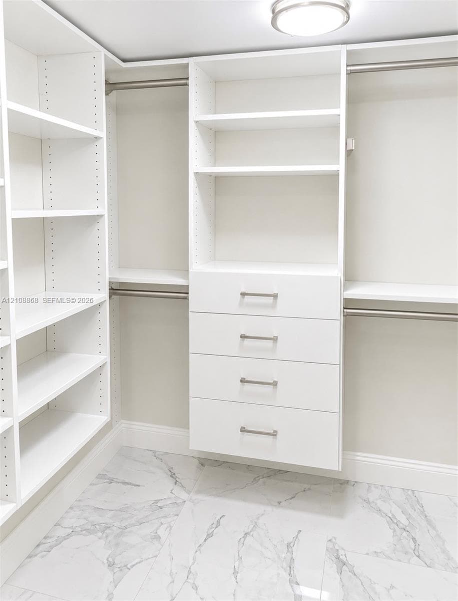 Walk-in closet featuring white built-in shelving, four soft-close drawers with metallic pulls, dual hanging rods, and large-format porcelain tile flooring