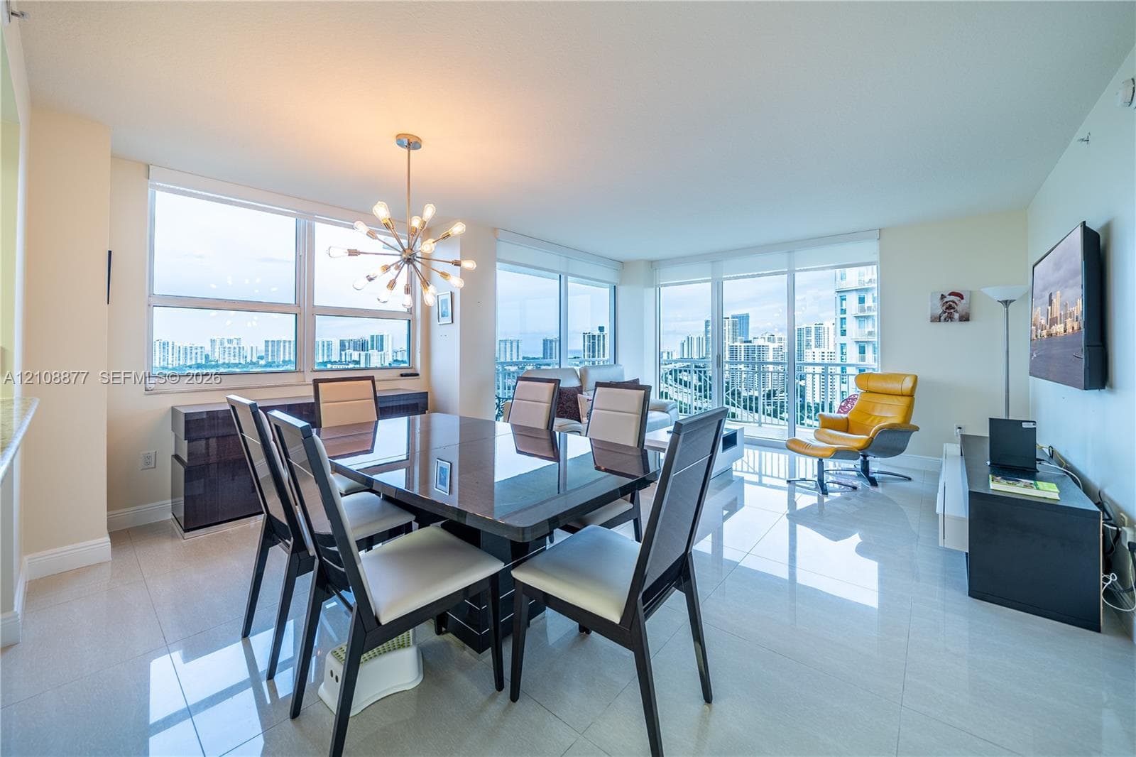 Open-concept living area featuring large floor-to-ceiling windows, polished tile flooring, a geometric multi-bulb chandelier, and access to a glass-railed balcony