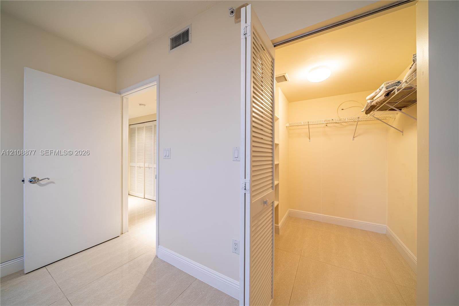 Walk-in closet featuring louvered bi-fold doors, white shelving units, and wire racking, complemented by large-format tile flooring and neutral wall tones