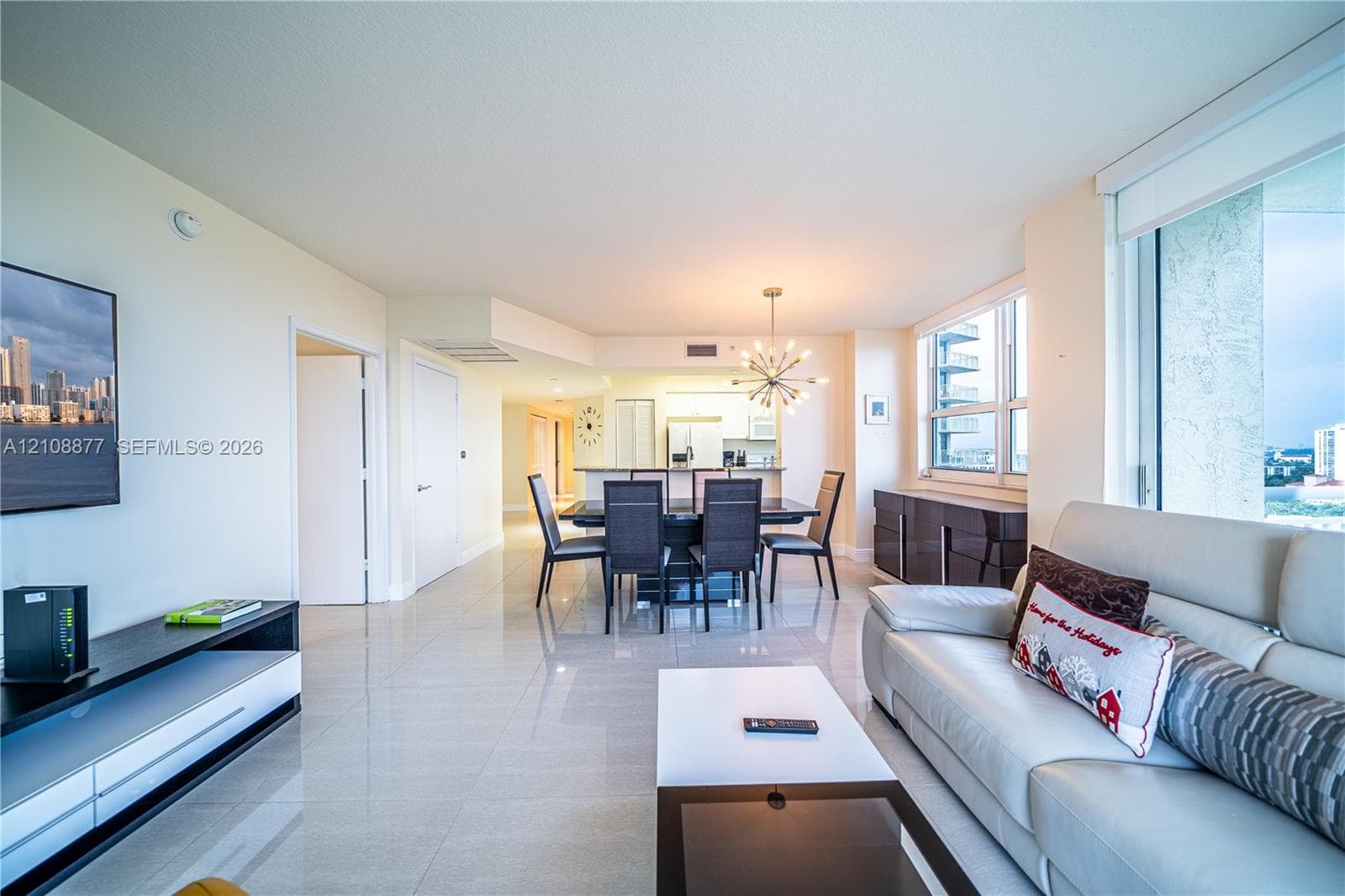 Open-concept living area featuring light-toned tile flooring, large windows, recessed ceiling lighting, and a multi-arm modern chandelier
