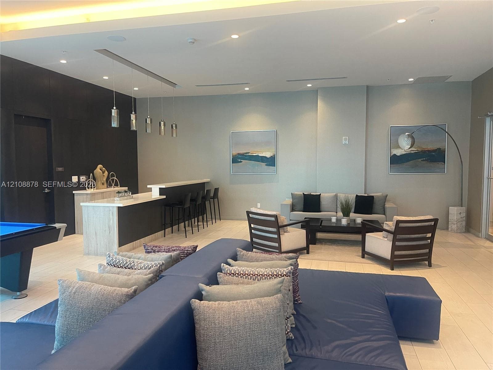 Open-concept interior featuring light-toned tile flooring, recessed ceiling lighting with perimeter cove illumination, a multi-arm modern chandelier, and a built-in bar with stone-surfaced countertops and hanging pendant lights