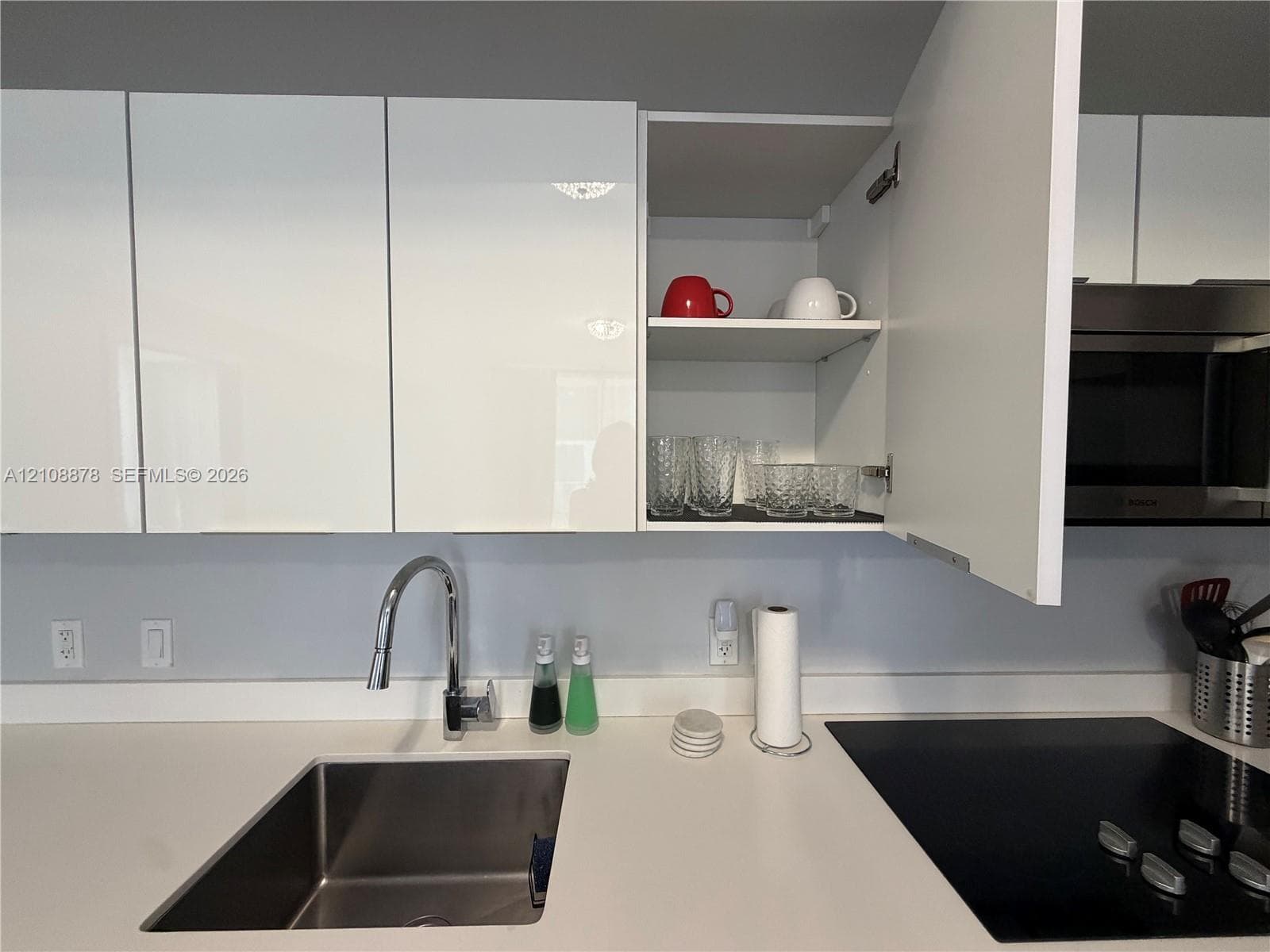 Glossy white flat-panel cabinetry, solid-surface countertop, undermount stainless steel sink, chrome pull-down faucet, and black electric cooktop