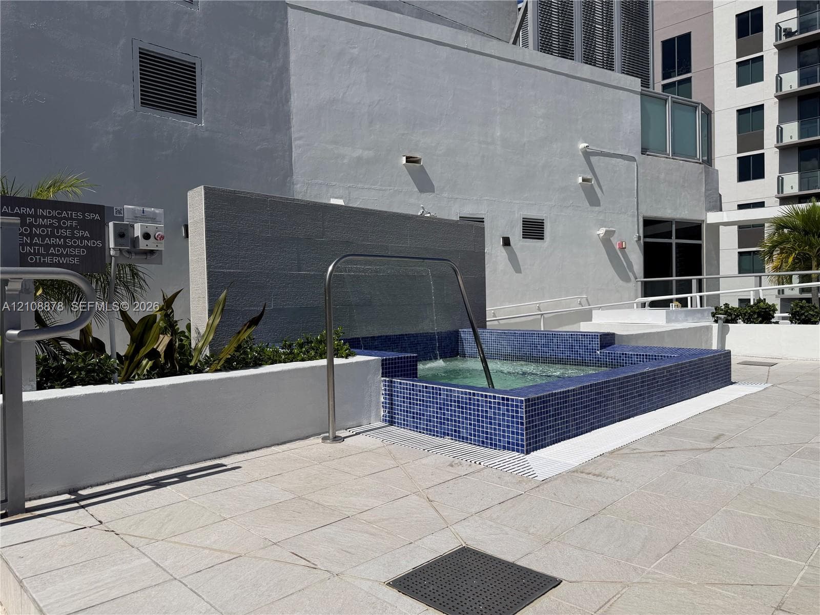 Raised square pool featuring cobalt blue glass tile finish, a stone-texture water feature wall, integrated tiered seating, and stainless steel handrail