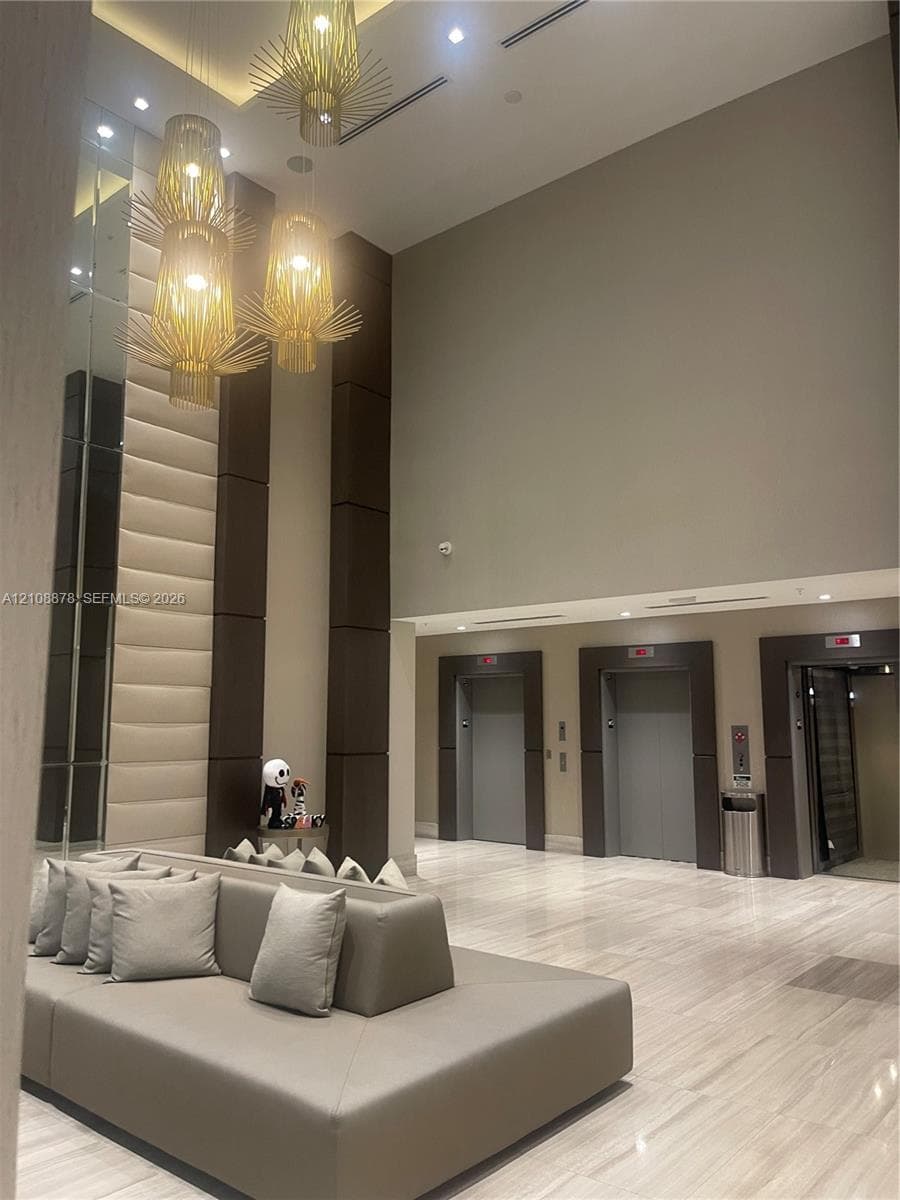 Double-height ceiling lobby featuring stone flooring, decorative metallic pendant lighting, floor-to-ceiling upholstered wall paneling, and multiple elevator access points