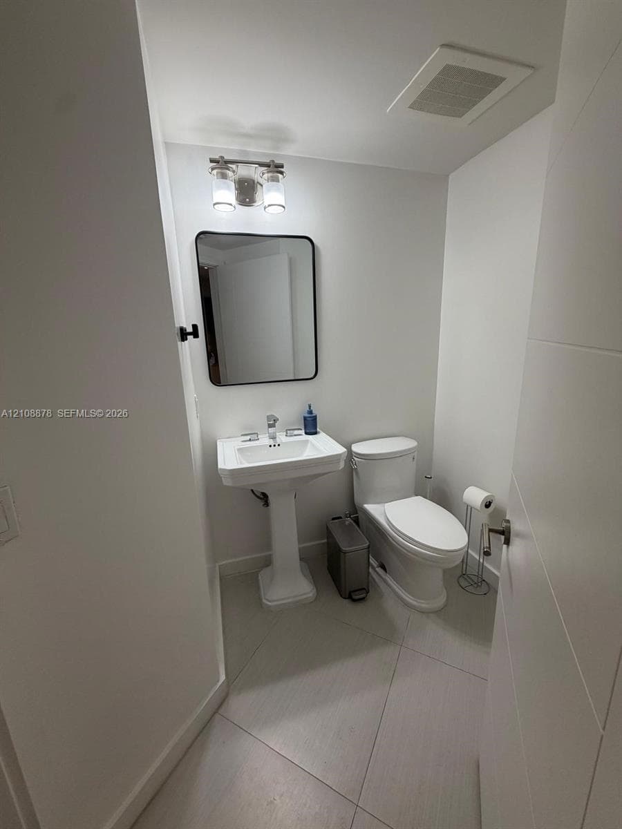 Pedestal sink, rectangular wall-mounted mirror with a dark frame, two-light fixture, and light-toned tile flooring