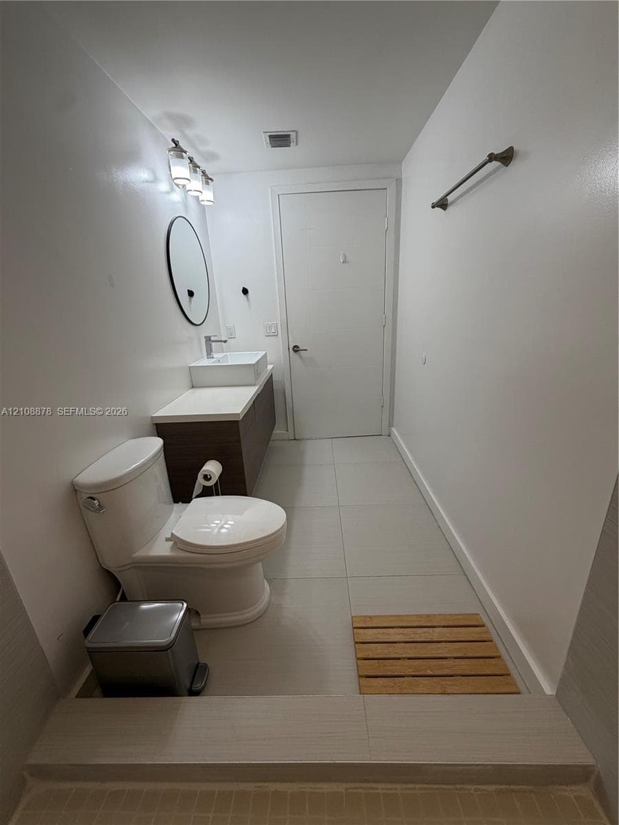 Floating dark wood-finish vanity with a white rectangular vessel sink, topped with a solid surface counter, circular gold-rimmed mirror, and large-format neutral floor tile