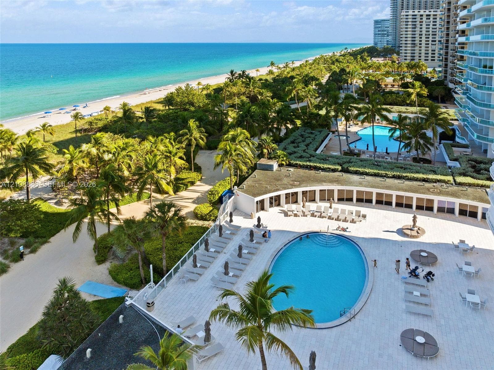 Aerial perspective featuring a direct oceanfront location, a large swimming pool, expansive paved sun terrace, manicured palm landscaping, and sandy beach access