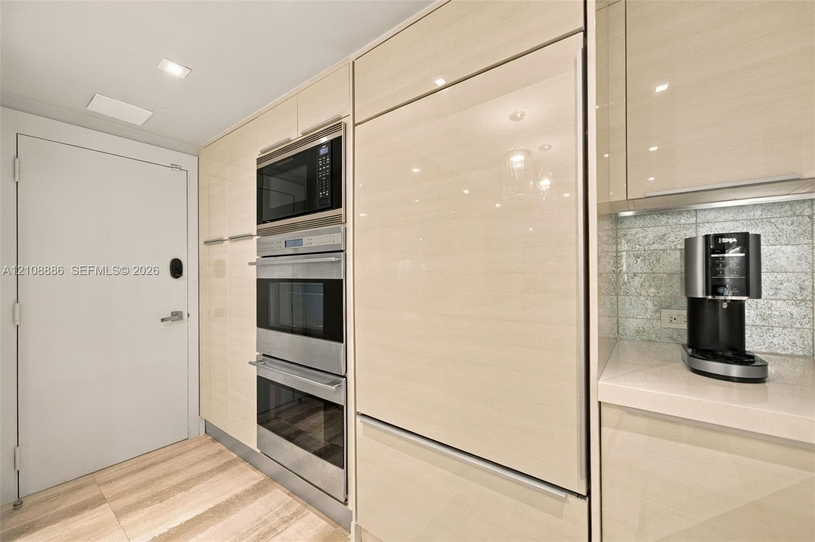 Sleek kitchen cabinetry with high-gloss finish, stainless steel wall-mounted microwave and double oven, integrated refrigerator panel, stone countertops, and a textured tile backsplash