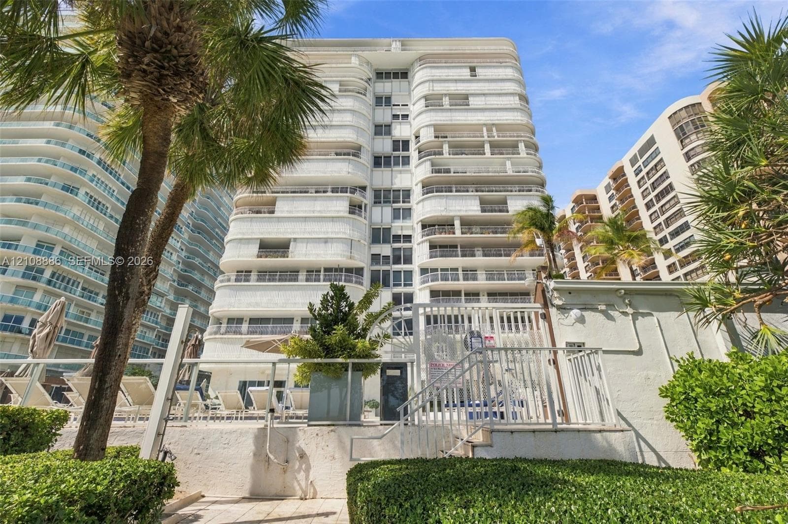 High-rise exterior featuring curved structural balconies, white facade, and integrated metal railings, set above a pool deck with palm landscaping and perimeter fencing