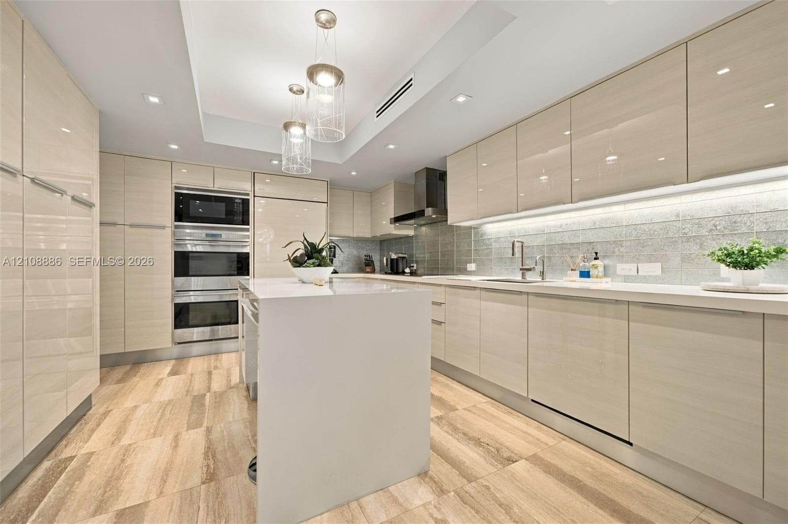 Contemporary kitchen featuring high-gloss cabinetry, waterfall edge island, stone tile flooring, stainless steel wall-mounted double oven, and recessed tray ceiling lighting