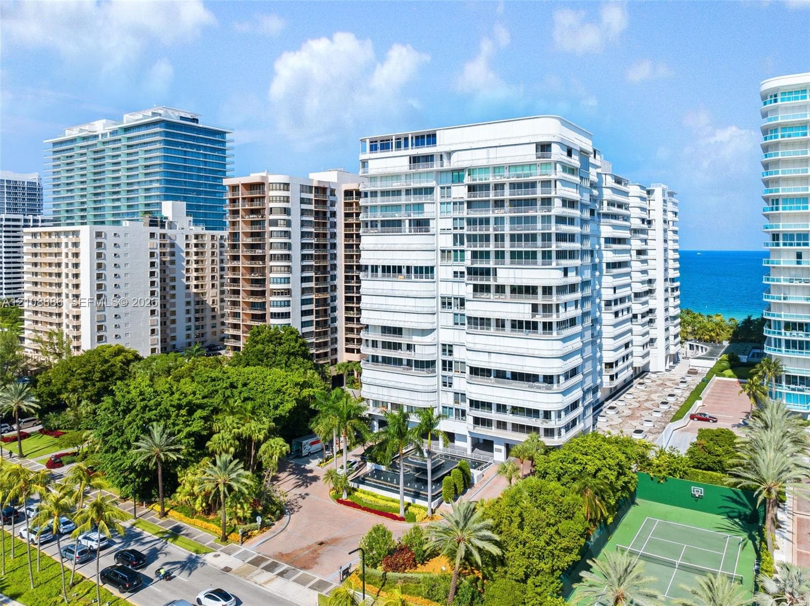 Mid-rise building featuring a white facade, private balconies, an on-site tennis court, landscaped grounds with mature palm trees, and water frontage