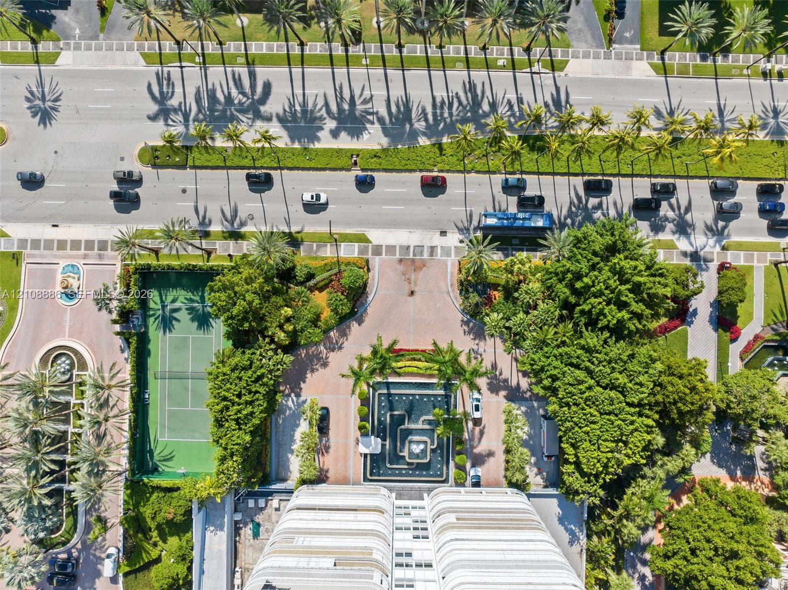Aerial perspective featuring a tennis court, stone-paved entry drive, central water feature, manicured landscaping, and mature palm trees