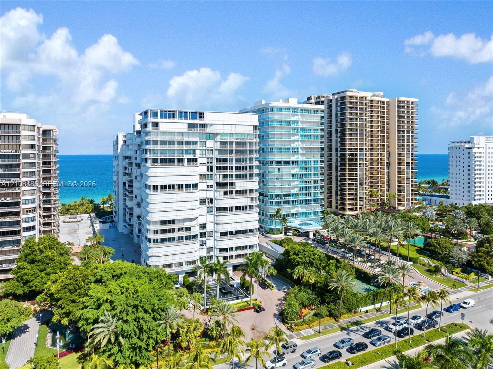 High-rise residential exterior featuring glass-railed private balconies, white facade detailing, and lush landscaped grounds with mature palm trees