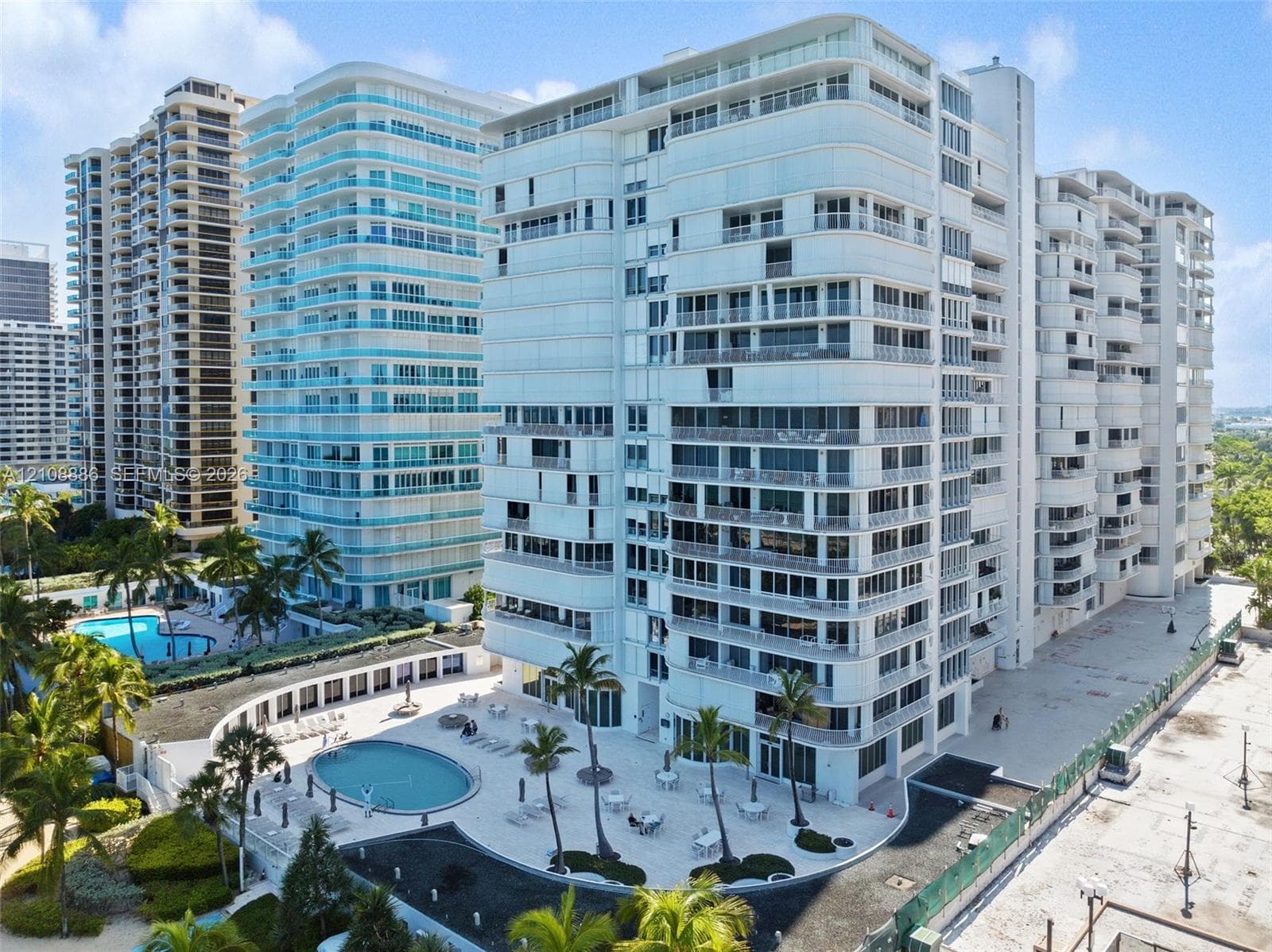 High-rise residential exterior featuring wrap-around balconies, a landscaped pool deck with multiple swimming pools, and palm tree-lined paved terrace areas