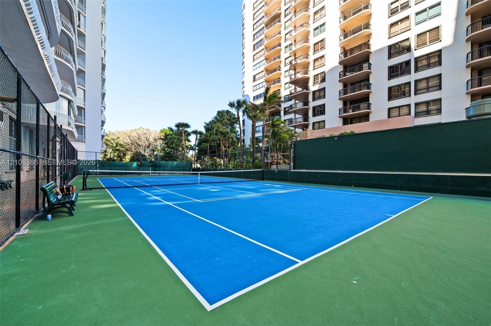 Outdoor tennis court featuring a blue playing surface, green perimeter zones, white court markings, and full-height chain-link fencing