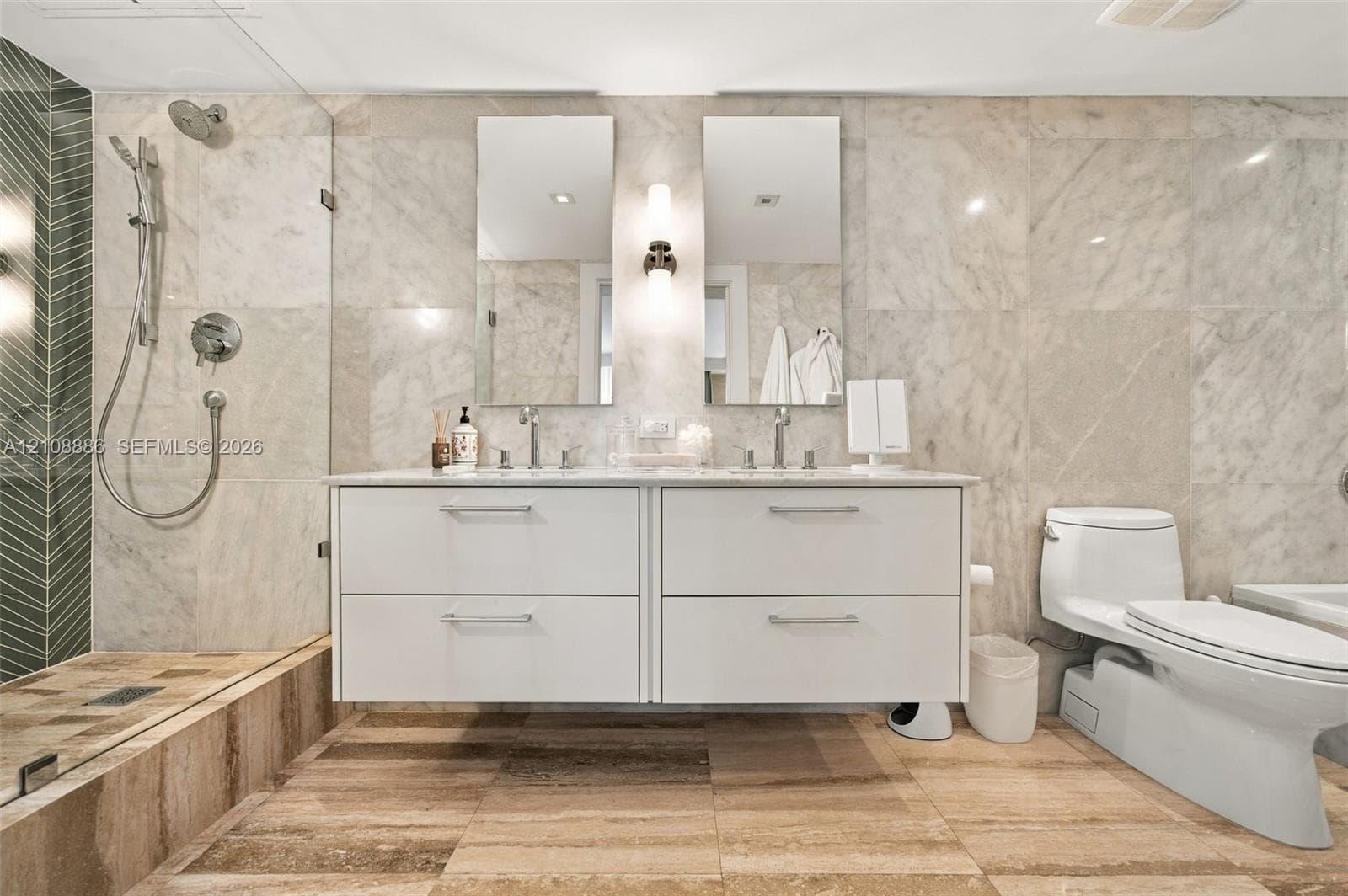 Floating white double vanity with dual undermount sinks, stone wall tiling, glass-enclosed walk-in shower with integrated hand-held sprayer, and tile flooring