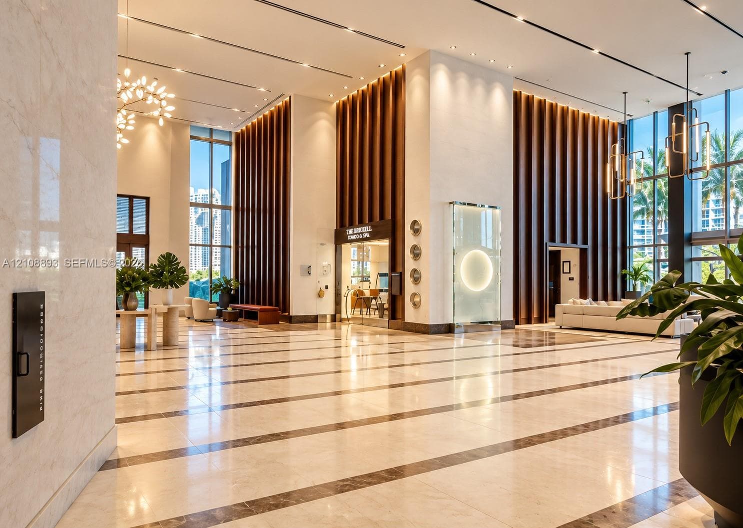 Grand lobby featuring high ceilings, floor-to-ceiling glass windows, polished stone flooring with inlaid borders, decorative vertical wood-slat wall accents, and custom multi-light chandelier fixtures