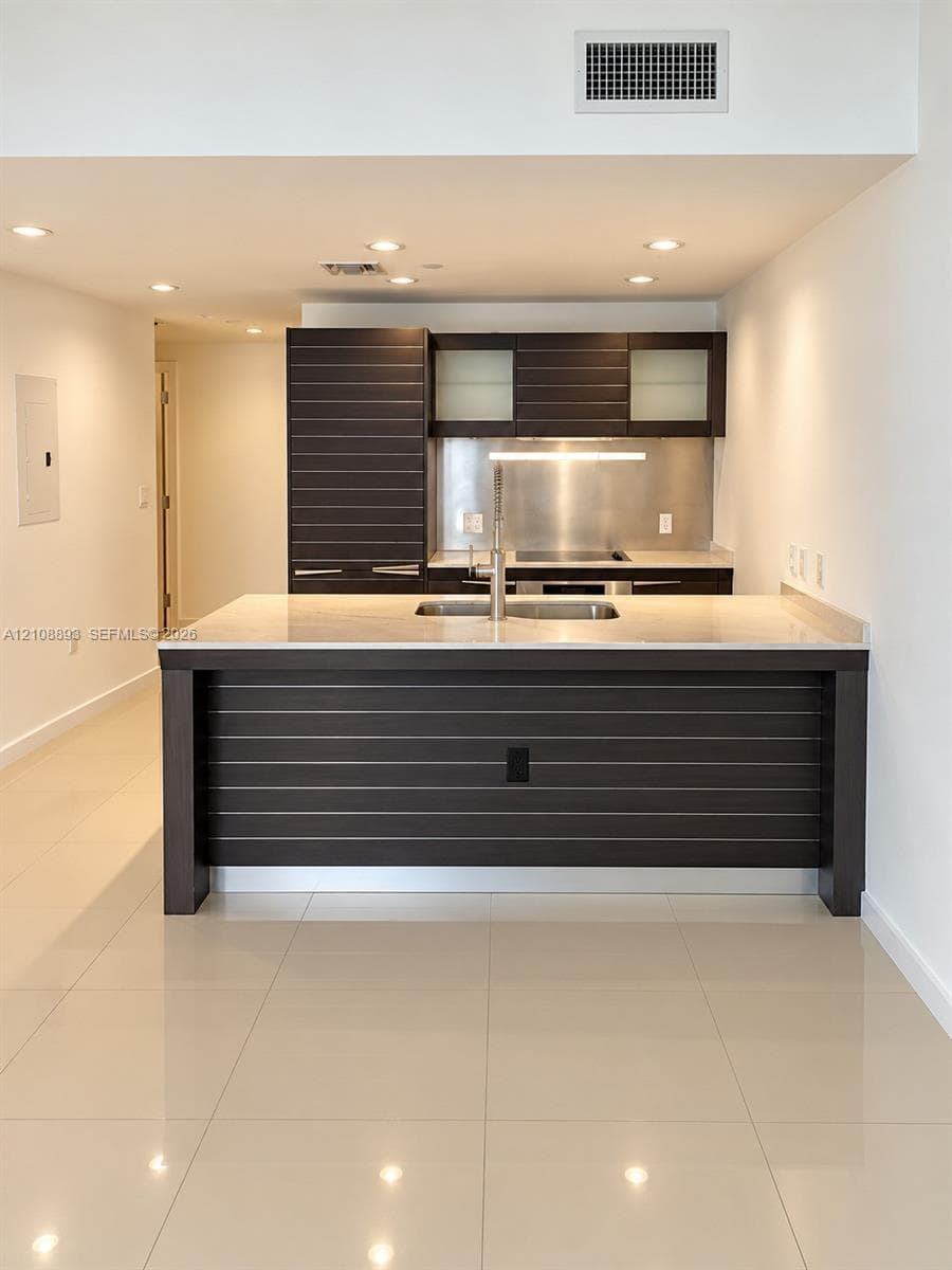 Kitchen featuring dark wood-finish cabinetry with horizontal slat detailing, stone-surface island countertop, integrated stainless steel backsplash, and polished tile flooring