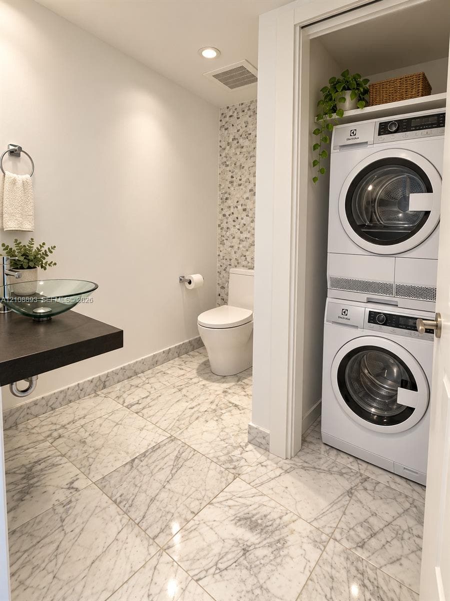 Full-bathroom featuring large-format stone-patterned floor tile, glass vessel sink with dark floating vanity, integrated laundry closet, and decorative stone-mosaic wall accent