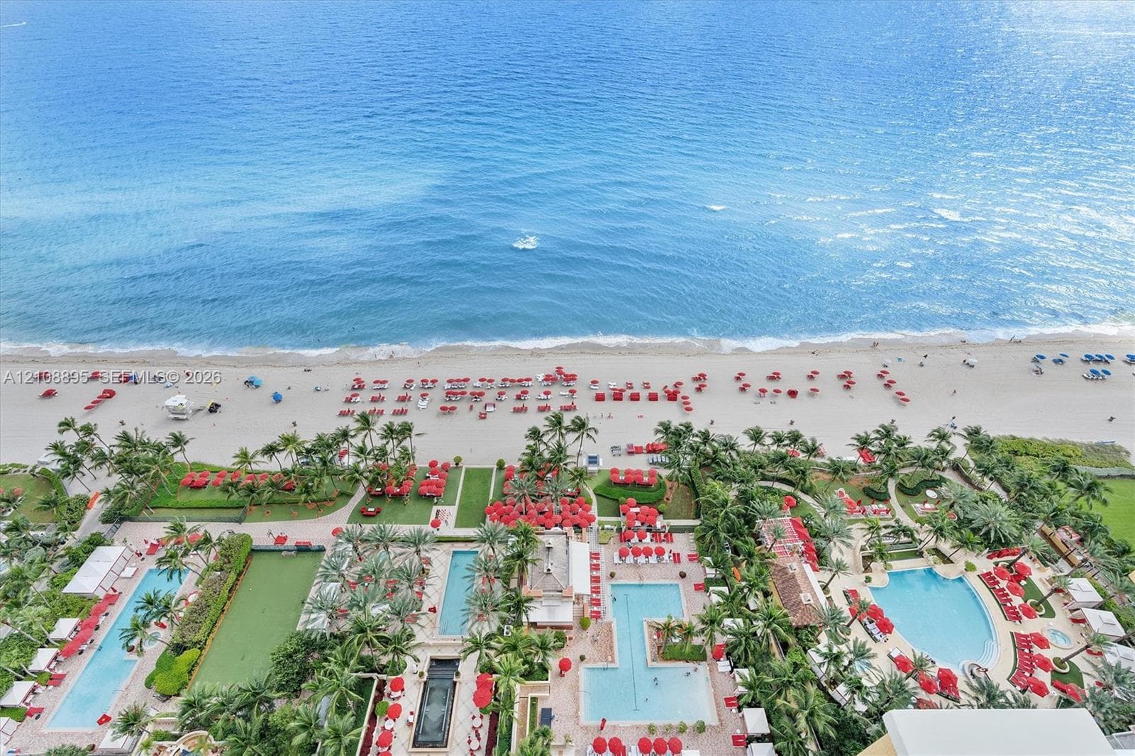 Aerial perspective of oceanfront property featuring multiple swimming pools, expansive paved sun decks, manicured palm landscaping, and direct access to the beach