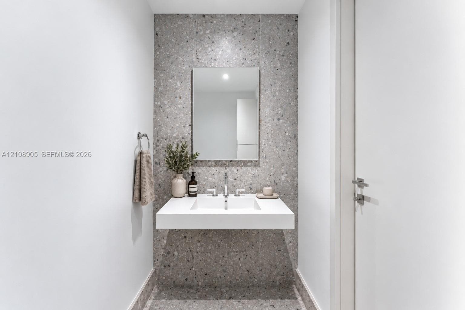 Floating vanity with a rectangular integrated sink, polished metal widespread faucet, rectangular wall-mounted mirror, and speckled stone-finish wall cladding