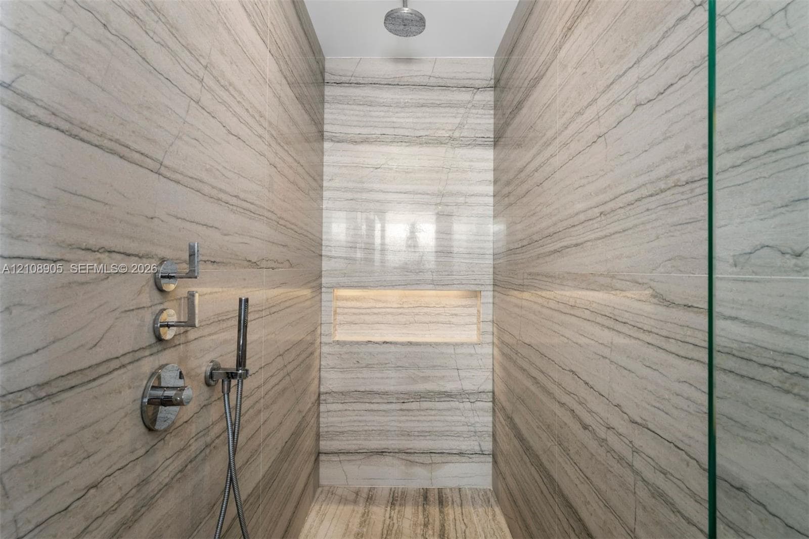 Walk-in shower featuring floor-to-ceiling stone-tiled walls, a built-in recessed niche, chrome wall-mounted fixtures, a handheld sprayer, and a ceiling-mounted rainfall showerhead