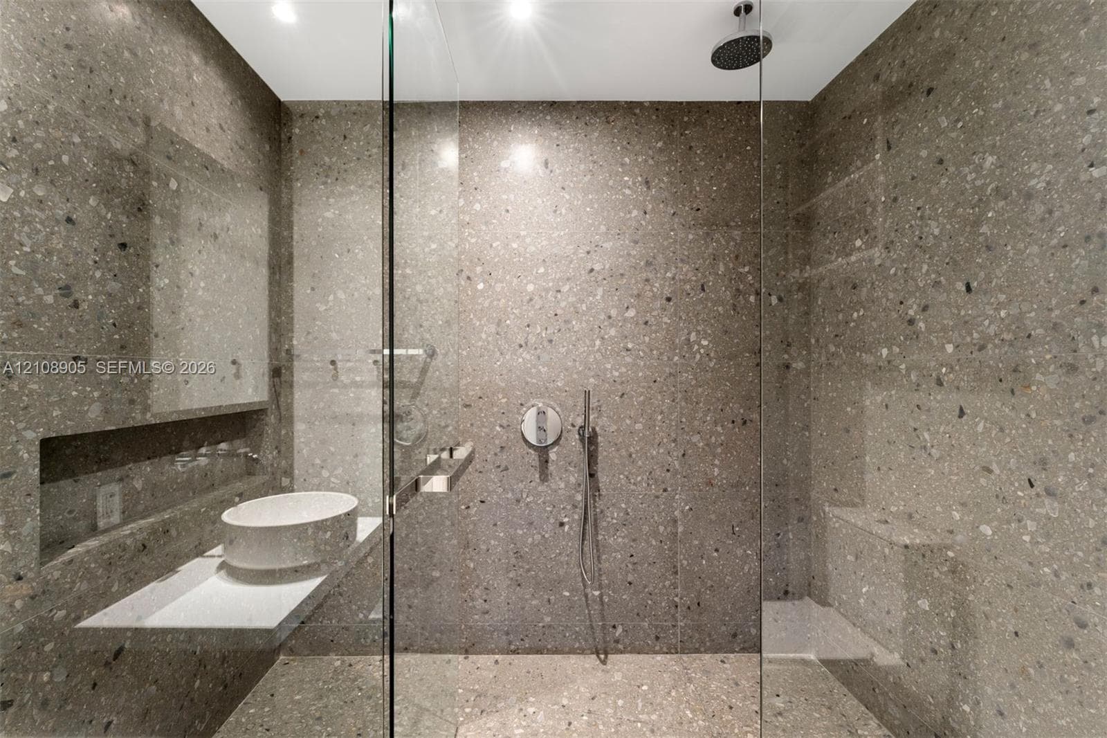 Full-height speckled stone wall finishes, glass-enclosed shower featuring a built-in bench, ceiling-mounted rainfall showerhead, and a floating vanity with a white vessel sink