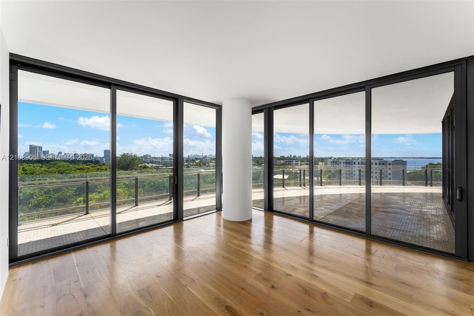 Floor-to-ceiling sliding glass walls, expansive wraparound balcony with metal railing, wide-plank wood-finish flooring, and structural cylindrical column
