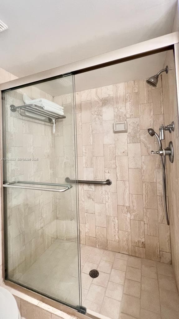 Walk-in shower featuring a sliding glass door, floor-to-ceiling neutral-toned tile, wall-mounted shower head with adjustable handheld attachment, and stainless steel grab bars