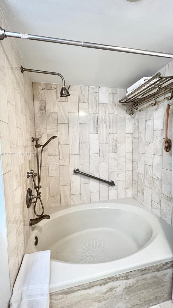 Oval soaking tub featuring a metal finish handheld shower head and rainfall fixture, offset by floor-to-ceiling neutral tile surround, a wall-mounted grab bar, and an integrated metal storage rack
