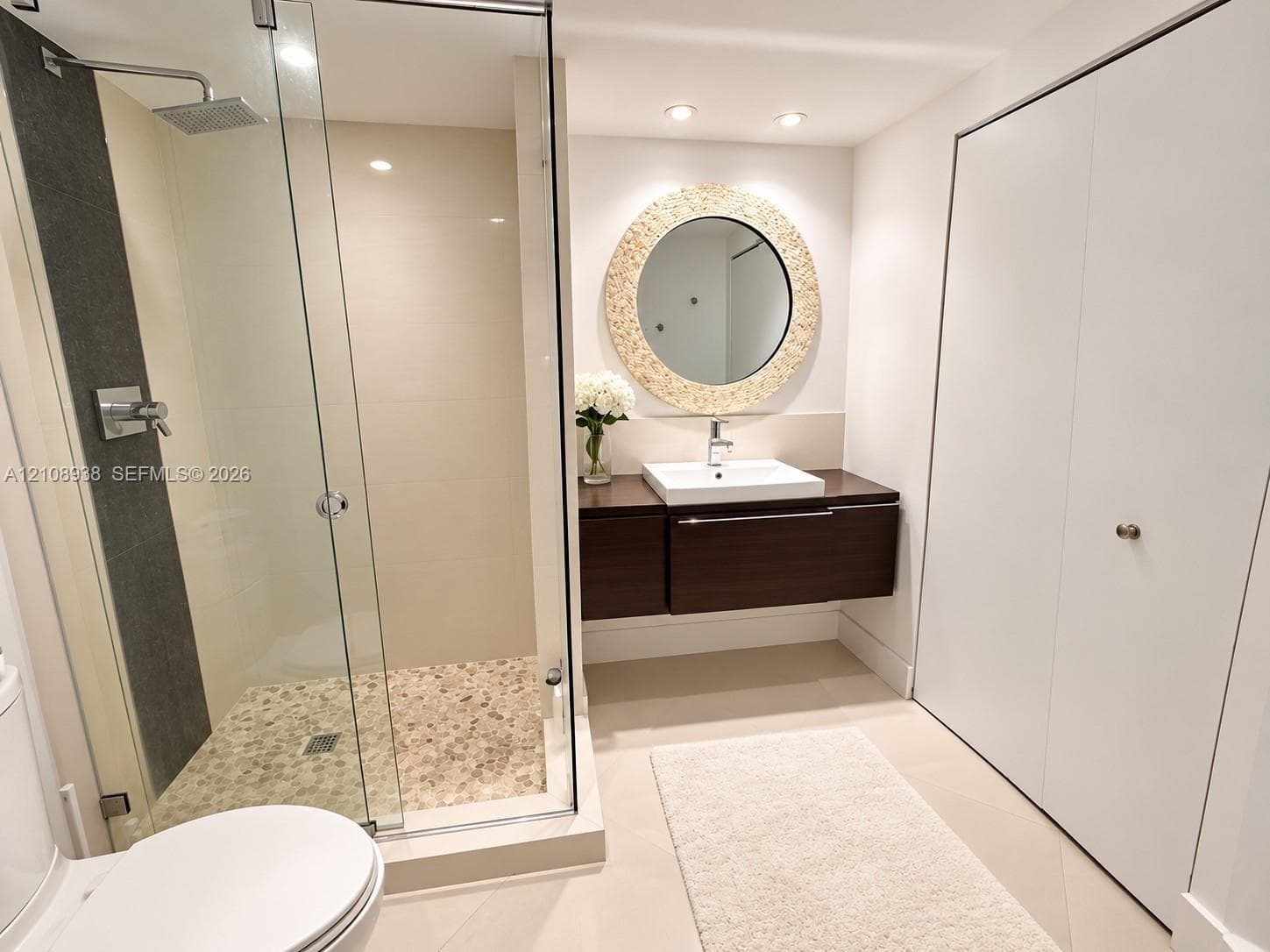 Floating dark wood-finish vanity with a rectangular vessel sink, glass-enclosed shower featuring a stone pebble floor, wall-mounted rainfall showerhead, and circular decorative mirror