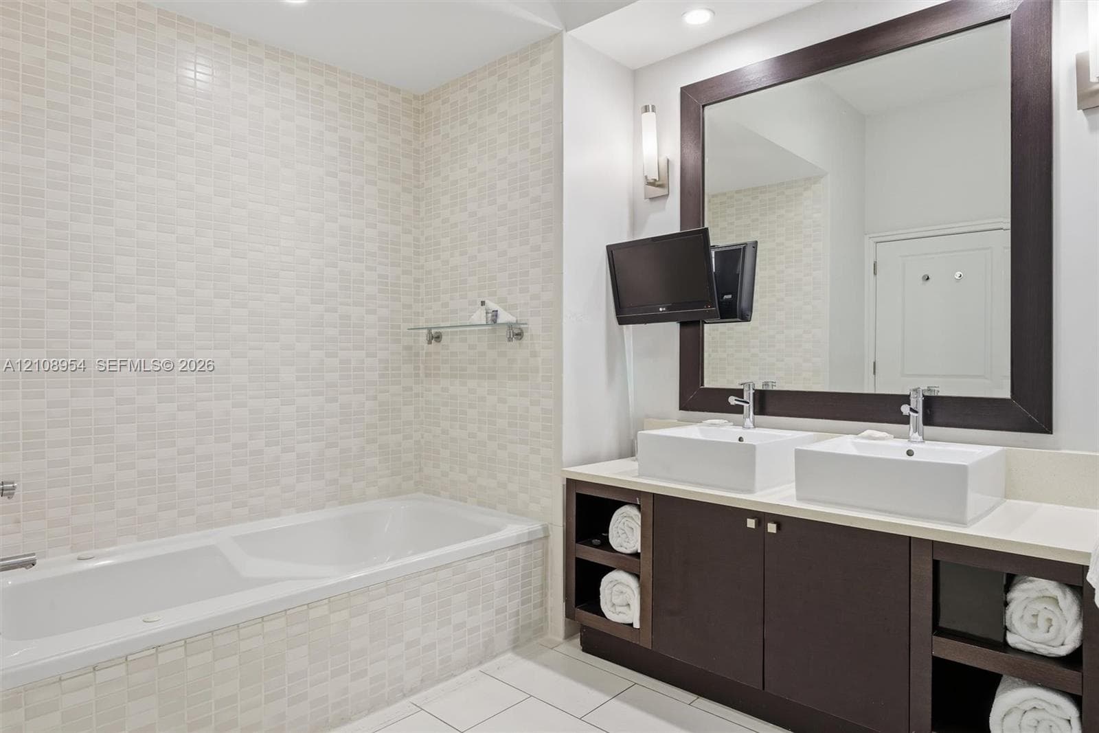 Dual vanity with rectangular vessel sinks, quartz-finish countertop, dark-toned cabinetry with open shelving, framed mirror, and tiled shower surround with built-in glass shelving