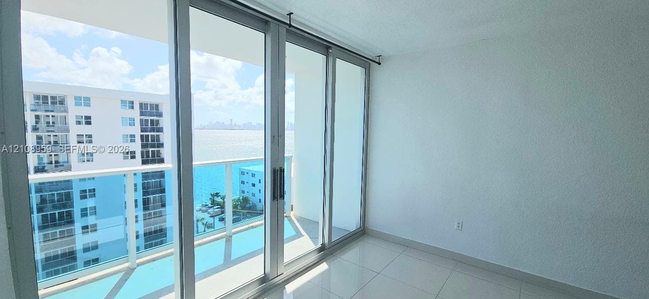 Floor-to-ceiling glass sliding doors, private balcony with glass railing, large-format tile flooring, and water backdrop
