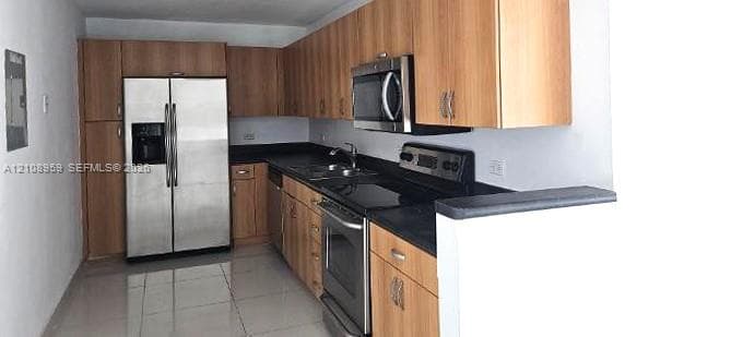 Kitchen featuring cabinetry with a wood finish, dark solid-surface countertops, stainless steel side-by-side refrigerator, stainless steel range and over-the-range microwave, and light-toned tile flooring