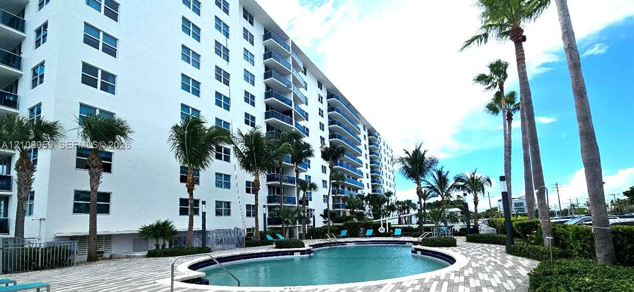 Multi-story exterior featuring private balconies with glass railings, a curved swimming pool, and stone-paver decking surrounded by palm trees