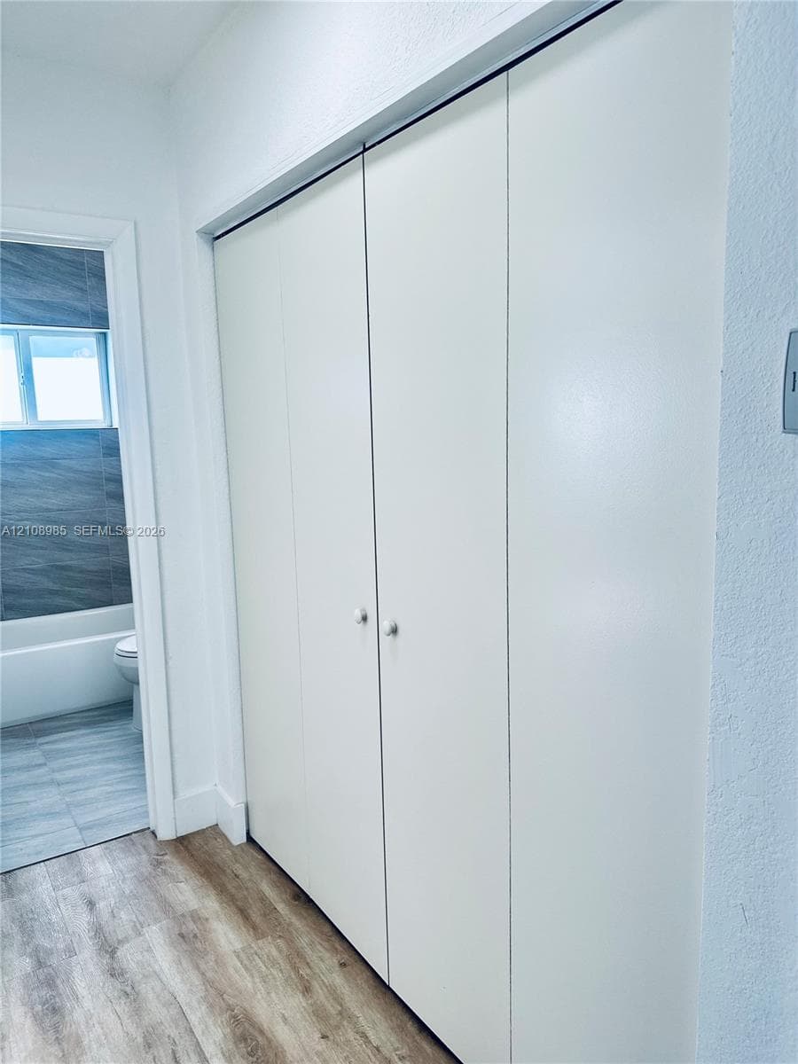 Full-height bi-fold closet doors with round knobs, wood-finish flooring, and a glimpse of a bathroom featuring tile flooring and a tiled tub surround