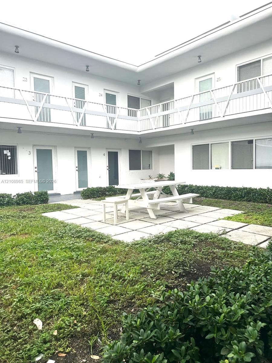 Courtyard exterior featuring a two-story building with white stucco siding, metal balcony railings, a central concrete paver patio, and low-lying perimeter shrubbery