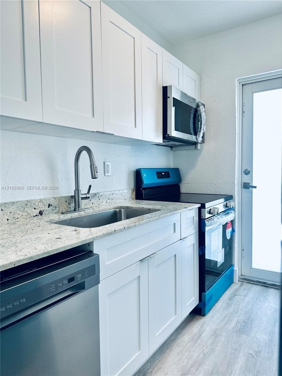 Kitchen featuring white shaker-style cabinetry, light-toned stone countertops, stainless steel appliances, a pull-down faucet, and wood-finish flooring