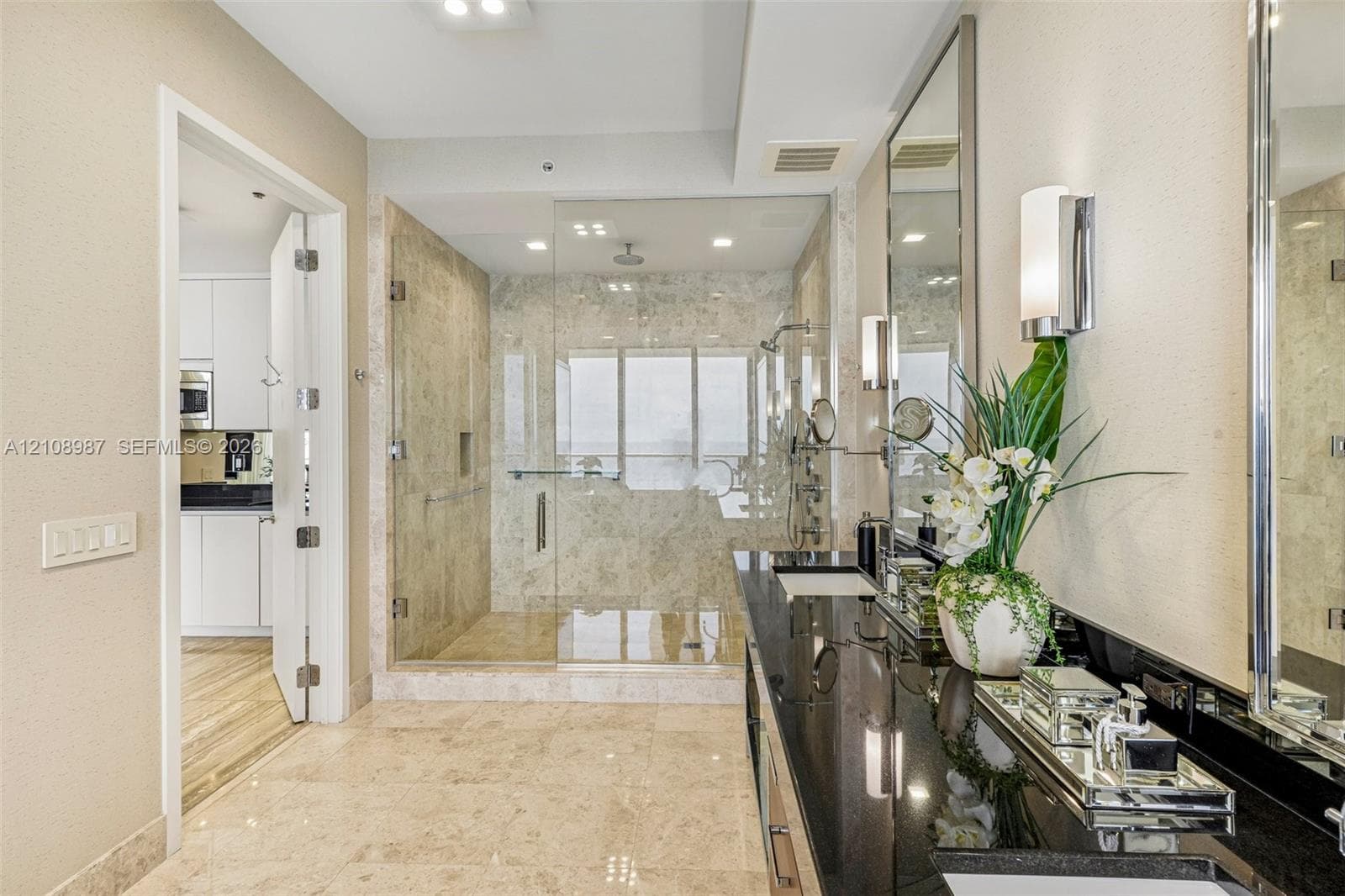 Bathroom featuring stone tile flooring, a glass-enclosed walk-in shower with stone surround, a dual vanity with a dark countertop, and full-length mirrors with integrated wall-mounted lighting