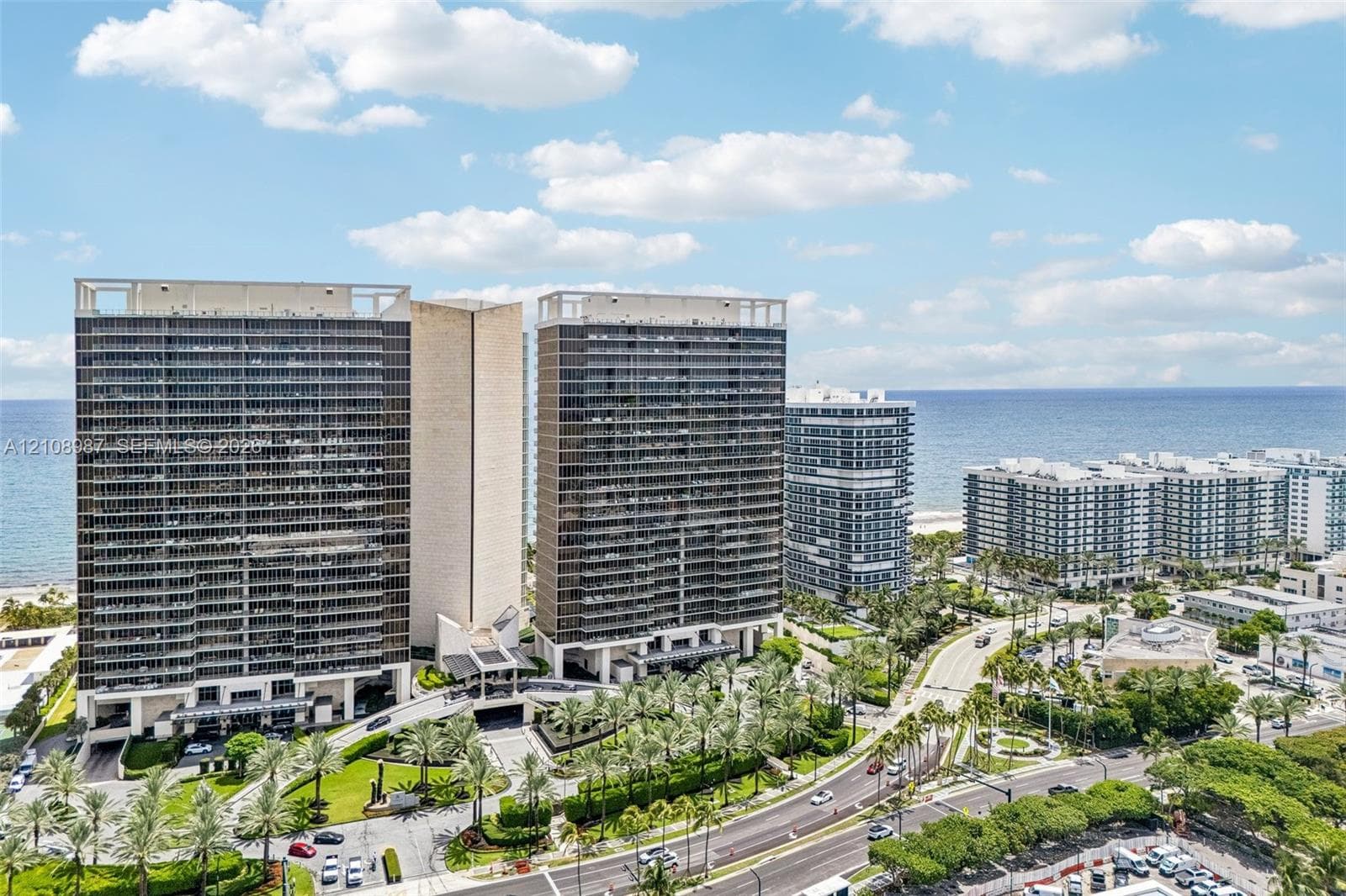 High-rise residential towers featuring balconies, glass-paneled facades, integrated covered ground-level parking, landscaped courtyards with palm trees, and direct oceanfront positioning